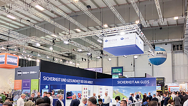 Besucherinnen und Besucher auf einer Fachmesse vor einem Messestand mit der Aufschrift „Sicherheit am Gleis“.