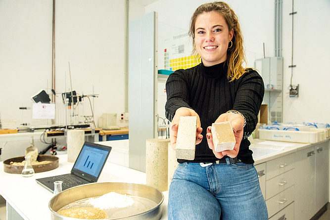 Wissenschaftliche Mitarbeiterin Pia Gebken zeigt erste Bindemittelversuche Eine junge Frau hält in einem Labor zwei Betonquader in der Hand, Neben ihr steht eine Schale mit einem weißen Pulver, Sand und Beton-Rezyklat.