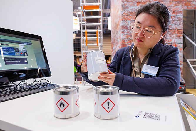 Eine Mitarbeiterin der BG BAU zeigt auf dem Messestand der BG BAU drei Dosen mit Gefahrstoffkennzeichnung.
