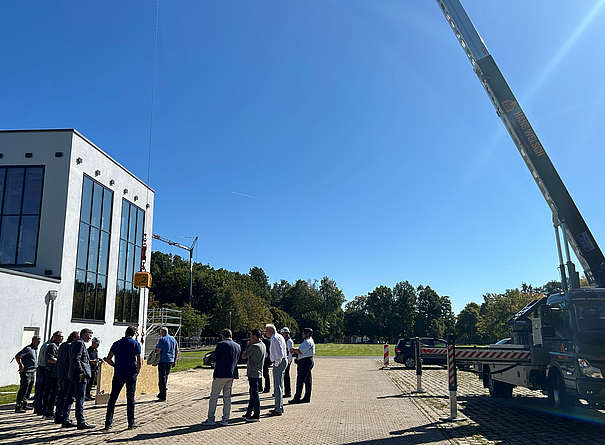 Eine Gruppe von Personen hat sich im Halbkreis vor einem modernen Gebäude versammelt und schaut auf eine Holzplatte, die an einem Kranseil hängt. 