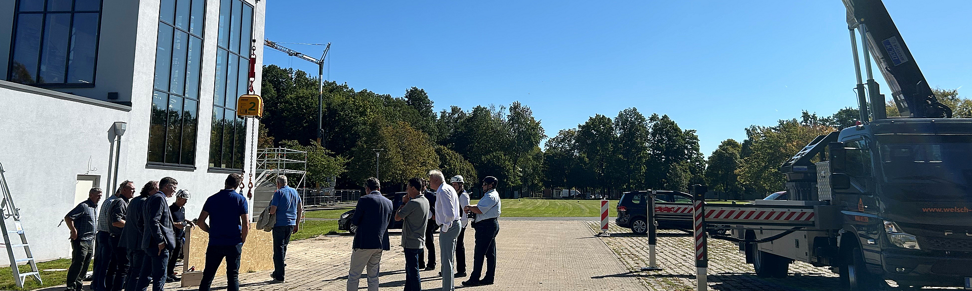 Eine Gruppe von Personen hat sich im Halbkreis vor einem modernen Gebäude versammelt und schaut auf eine Holzplatte, die an einem Kranseil hängt. 