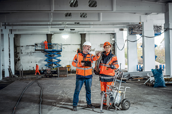 Zwei Personen mit Schutzkleidung schauen sich die Sanierungsarbeiten in der Halle an.