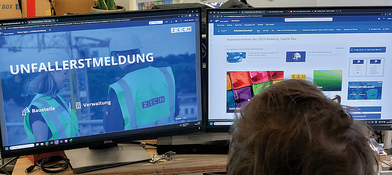 Eine Person sitzt vor zwei Monitoren. Auf dem linken Bildschirm ist die Startseite der digitalen Unfallerstmeldung geöffnet, 