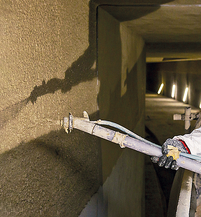 Ein Beschäftigter spritzt den Brandschutzputz auf die Tunnelwand. Er trägt einen Schutzanzug und Arbeitshandschuhe.