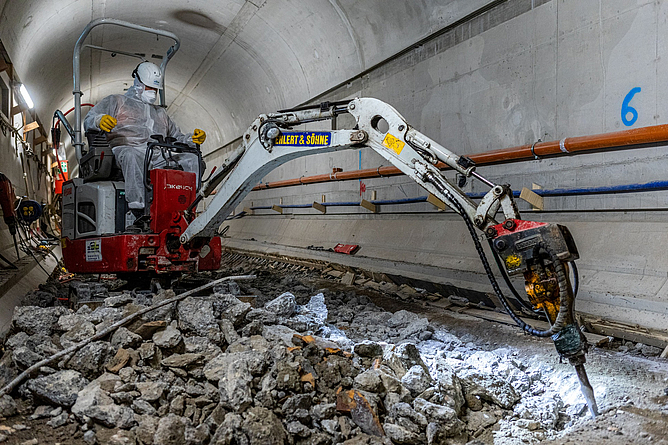 Person mit Vollschutzausstattung sitzt in einer schmalen Tunnelröhre auf einem maschinenbetriebenen Abbruchgerät. 