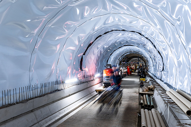 Sanierung Alter Elbtunnel, Einbau einer Flächendränage und einer Abdichtung oberhalb des Sockelbereichs Mit heller Folie abgedichteter Tunnel, in dessen unterem Bereich ein Betonrand mit Bewehrungseisen ist. Rechts neben dem Betonrand liegen Rohrteile.