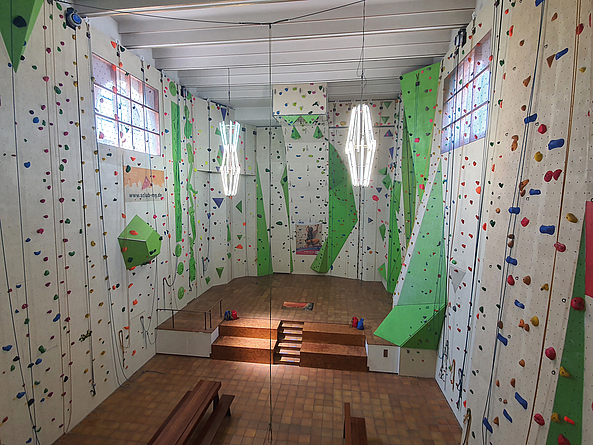 Innenraum einer Kletterhalle mit hohen Wänden, bunten Griffen und mehreren Seilen; in der Mitte eine erhöhte Plattform mit Treppe.