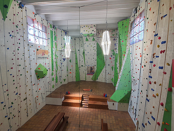 Innenraum einer Kletterhalle mit hohen Wänden, bunten Griffen und mehreren Seilen; in der Mitte eine erhöhte Plattform mit Treppe.