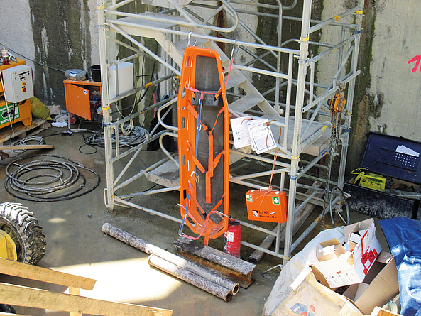 	Eine orange Trage, die senkrecht an einem Treppenturm hängt. Dieser befindet sich in einem Baufeld.