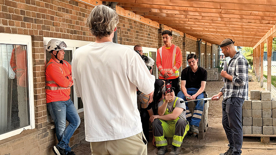 Mehrere Personen mit Warnwesten und Schutzhelmen nehmen auf einer Baustelle an einer Besprechung teil; einige stehen, andere sitzen.