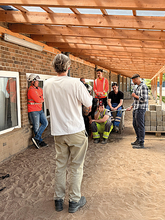 Mehrere Personen mit Warnwesten und Schutzhelmen nehmen auf einer Baustelle an einer Besprechung teil; einige stehen, andere sitzen.