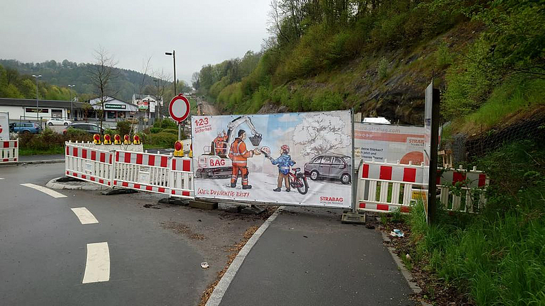 Abgesperrte Straße, am Absperrzaun ist ein comicartiges Plakat befestigt. 