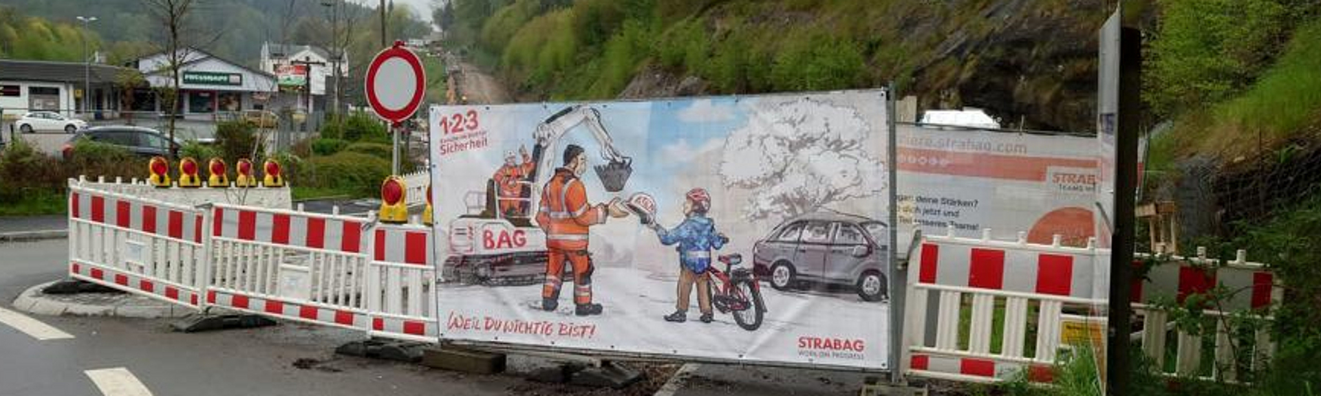 Abgesperrte Straße, am Absperrzaun ist ein comicartiges Plakat befestigt. 