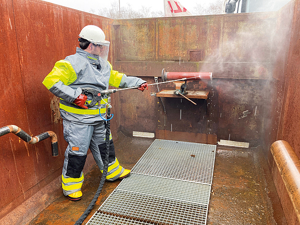 Mann in Schutzkleidung mit einer handgehaltenen Spritzeinrichtung und Hochdruckwasserstrahl. Zur Schutzkleidung gehören der Schutzhelm mit Gesichtsschutz und Gehörschutz, Schutzhandschuhe und Schutzstiefel.