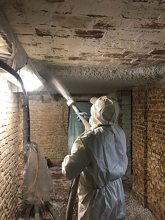 Spritzarbeiten unter Vollschutz Person mit Vollschutzkleidung spritzt in einem Kellerraum aus einer Pumpe etwas an die Decke.