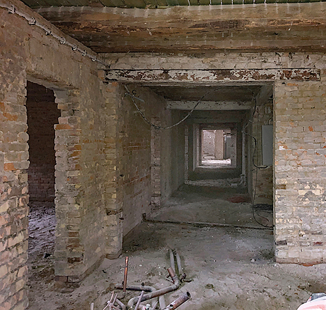 Ein Gebäudeflur im Rohbauzustand zeigt unverputzte Ziegelwände, offene Türdurchgänge und freiliegende Kabel. 