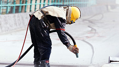 Beschäftigte oder Beschäftigter mit Persönlicher Schutzausrüstung, die oder der auf einer Brücke mit den Betonboden mit Strahlgut bearbeitet. 