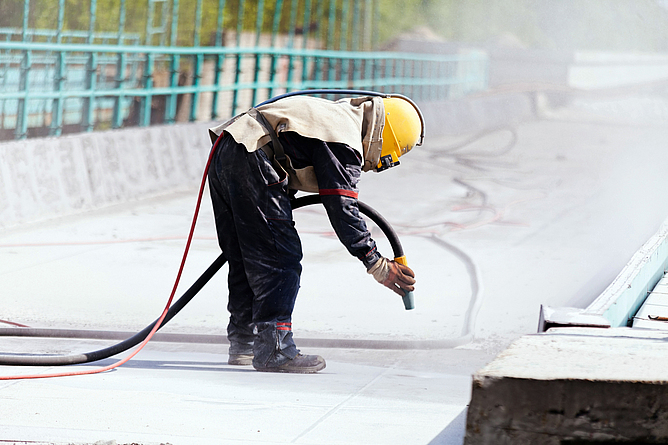 Beschäftigte oder Beschäftigter mit Persönlicher Schutzausrüstung, die oder der auf einer Brücke mit den Betonboden mit Strahlgut bearbeitet. 