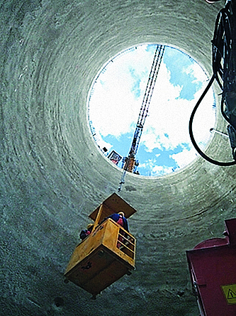 Blick von der Sohle einer Baugrube nach oben: Über einen Arbeitskorb, der mit Seilen befestigt an einem Kran hängt, werden Beschäftigte in die Tiefe hinab befördert.