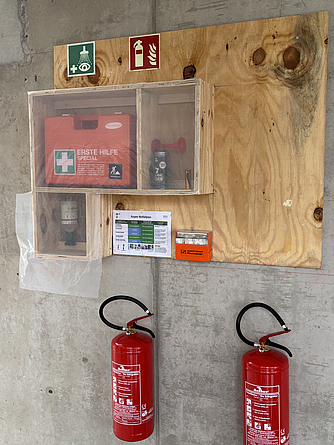 Auf einer Baustelle sind an einer Holztafel ein Erste-Hilfe-Kasten, Sicherheitszeichen, ein Feuerlöscher und Informationsblätter angebracht.