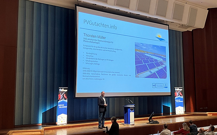 Vortrag Thorsten Müller auf dem Fachkongress für Absturzsicherheit 2023 Ein Mann hält auf einer Bühne einen Vortrag, hinter ihm ist eine Präsentation auf Leinwand zu sehen.