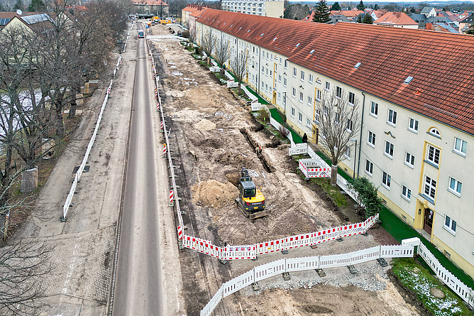 Mit Bauzäunen abgesperrtes Gebieten zwischen Wohngebäuden und einer Straße. Auf einem Baufeld steht ein Bagger.