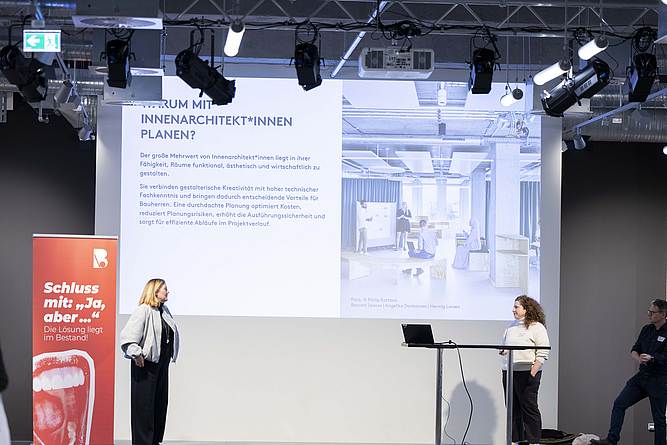 Podium mit zwei vortragenden Frauen vor Präsentation.