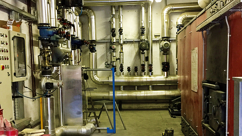 Die Wärmeenergie kommt vom benachbarten Holzbauunternehmen LignoAlp, zu sehen ist die Abgabestation. Abgabestation in einem hohen Raum mit vielen Metallrohren