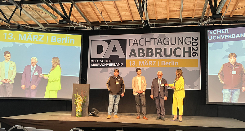 Drei Männer und eine Frau sind auf einem Podium. Im Hintergrund große Präsentationswände.