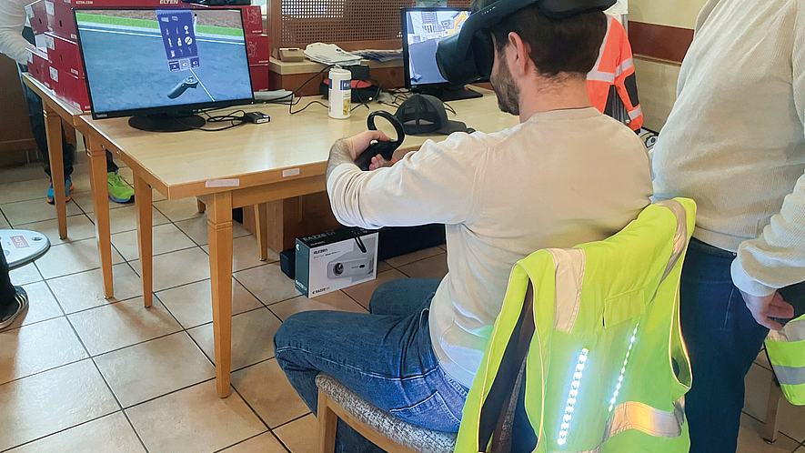 Person mit VR-Brille sitzt an einem Tisch und hält einen Controller vor zwei Monitoren.