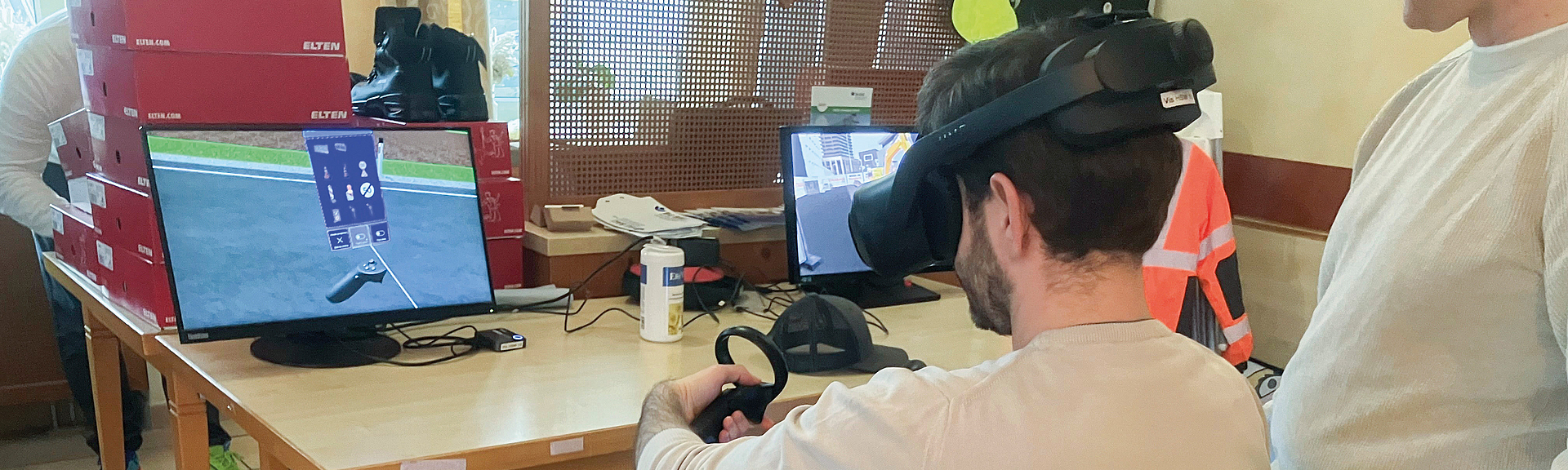 Person mit VR-Brille sitzt an einem Tisch und hält einen Controller vor zwei Monitoren.