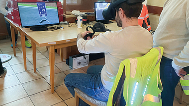 Person mit VR-Brille sitzt an einem Tisch und hält einen Controller vor zwei Monitoren.