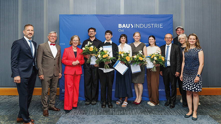 Gruppenfoto von elf Personen vor einer blauen Bauindustrie-Pressewand; vier Personen in der Mitte halten Blumensträuße und Urkunden.