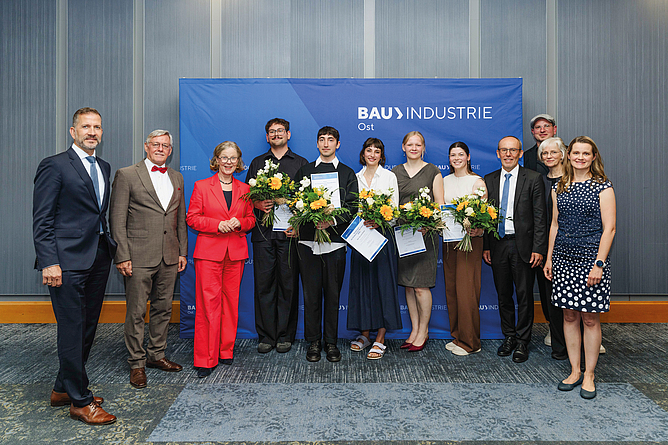 Gruppenfoto von elf Personen vor einer blauen Bauindustrie-Pressewand; vier Personen in der Mitte halten Blumensträuße und Urkunden.