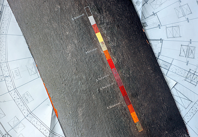 Ein alt wirkendes Metallstück mit einer Farbskala liegt auf einem Papierplan.