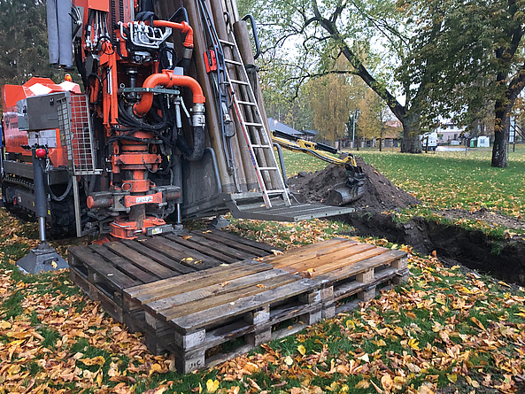 Auf einer mit Laub bedeckten Wiese steht ein Bohrgerät neben einer Baugrube. 