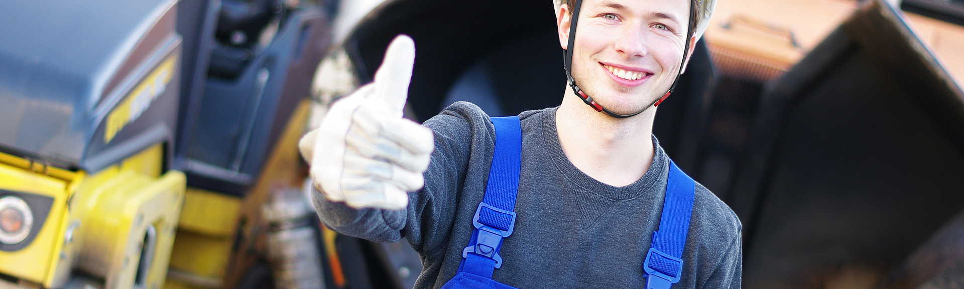 Ein junger Bauarbeiter mit Schutzhelm hält auf einer Baustelle lächelnd den Daumen hoch in die Kamera.
