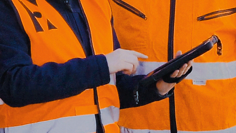 Drei Personen mit blauem Helm und Schutzweste, die vor einer Baumaschine stehen und auf ein Smartphone schauen.