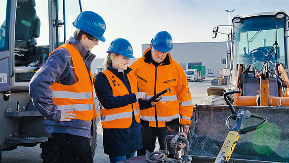 Bausituation mit Digitaltool Drei Personen mit blauem Helm und Schutzweste, die vor einer Baumaschine stehen und auf ein Smartphone schauen.