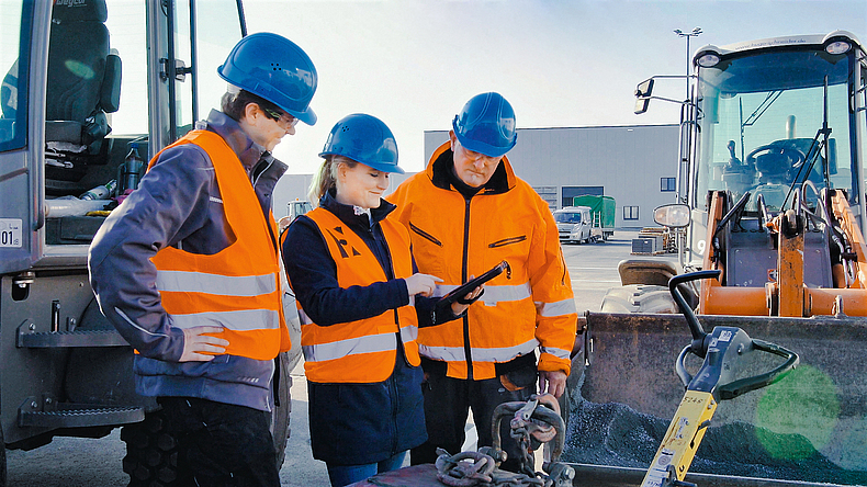 Drei Personen mit blauem Helm und Schutzweste, die vor einer Baumaschine stehen und auf ein Smartphone schauen.