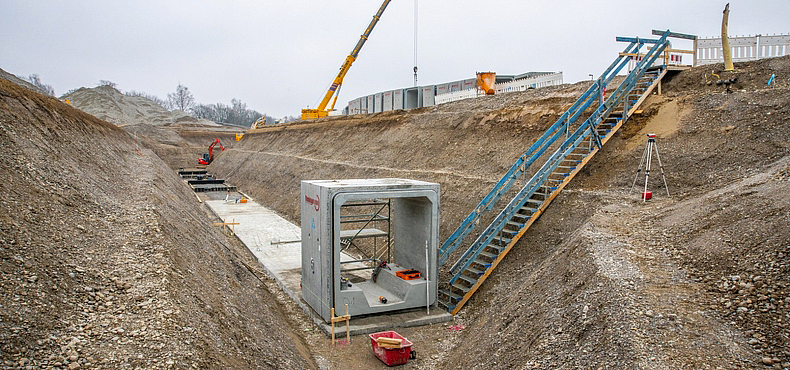 Längliche Baugrube mit einem aufgebauten Rechtprofil. In die Baugrube führt eine Bautreppe mit Geländer.