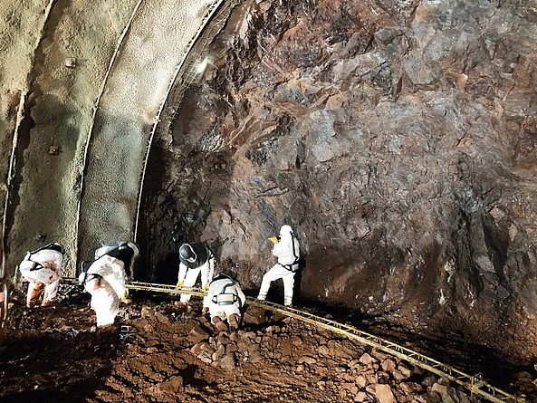 Im Brandbergtunnel arbeiten sechs Beschäftigte in persönlicher Schutzausrüstung (Schutzanzug, Helm, Atemschutzgeräte) an der Montage einer Transportschiene zum Abtransport des asbesthaltigen Abraums beim Tunnelvortrieb.