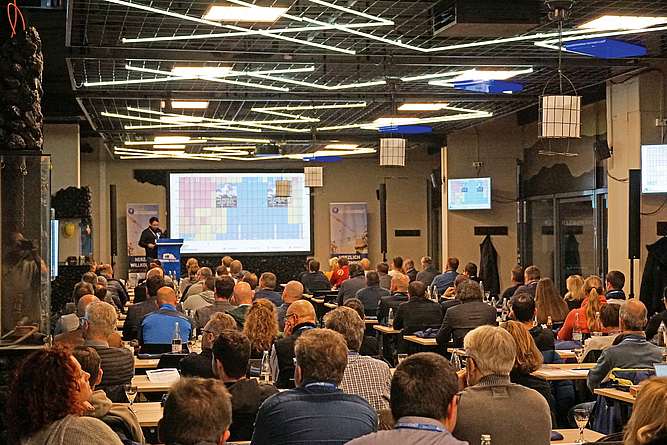 Vortragsraum mit an Tischen sitzenden Teilnehmenden. Vorne referiert David Auerbach an einem Pult.