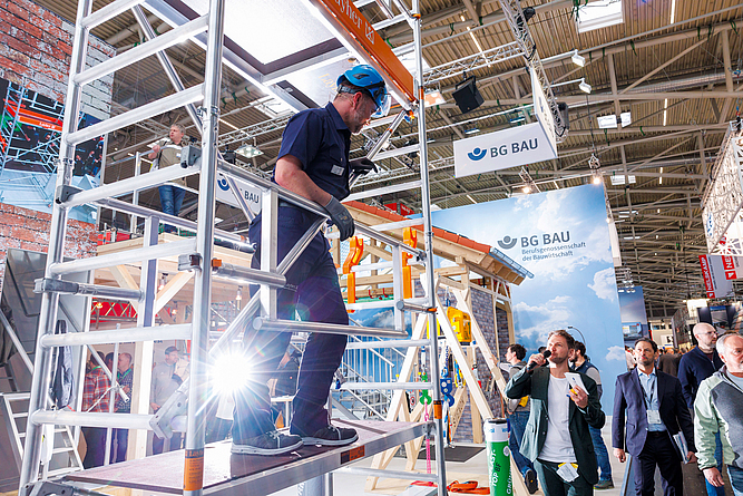 Stand der BG BAU auf der Messe BAU. Ein Mann steht auf einem Gerüst mit Seitenschutzgeländer. Messebesucher schauen zu, während ein Standbetreuer die Vorführung moderiert.