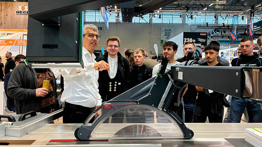 Gruppe von Männern steht auf einer Messe um eine Kreissäge; ein Mann erklärt und zeigt auf ein Bauteil.