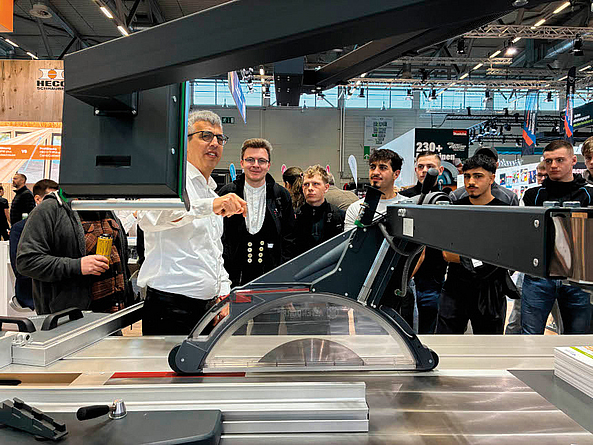Gruppe von Männern steht auf einer Messe um eine Kreissäge; ein Mann erklärt und zeigt auf ein Bauteil.