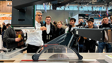 Gruppe von Männern steht auf einer Messe um eine Kreissäge; ein Mann erklärt und zeigt auf ein Bauteil.