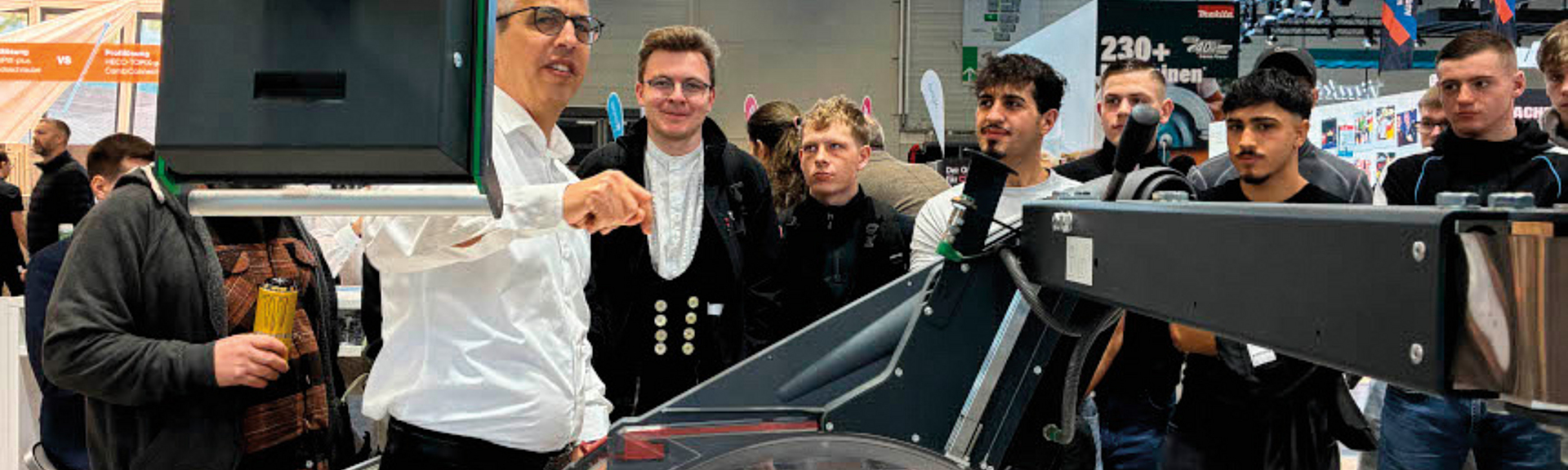 Gruppe von Männern steht auf einer Messe um eine Kreissäge; ein Mann erklärt und zeigt auf ein Bauteil.