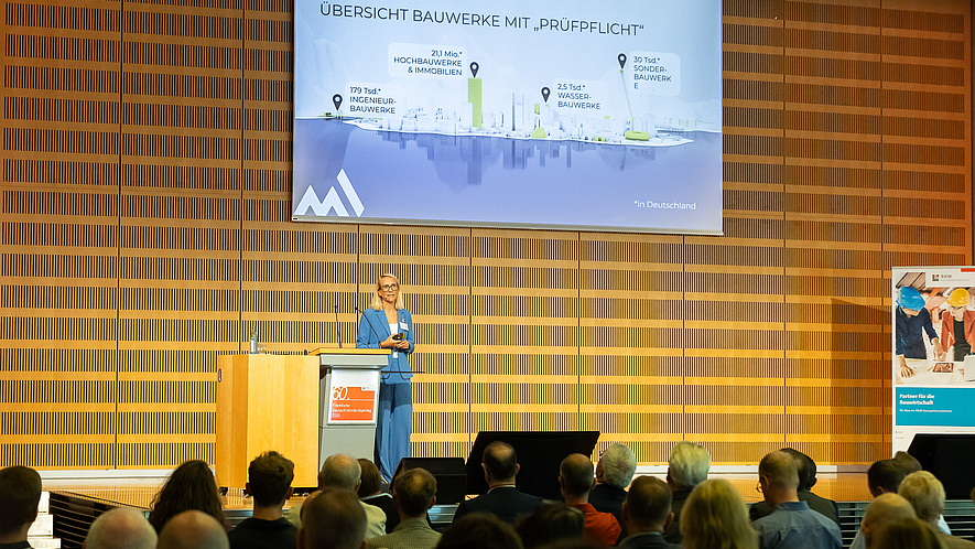 Frau steht auf einer Bühne und hält einen Vortrag vor Publikum, im Hintergrund eine Präsentationsfolie mit dem Titel „Übersicht Bauwerke mit Prüfpflicht“.