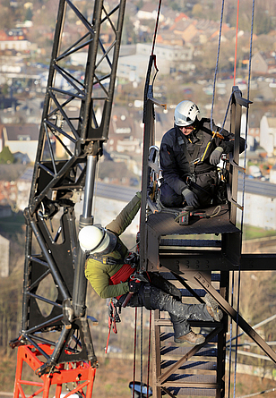 Zwei Personen mit Schutzhelm und seilgesichert am Kran arbeiten hoch oben am Gasometer